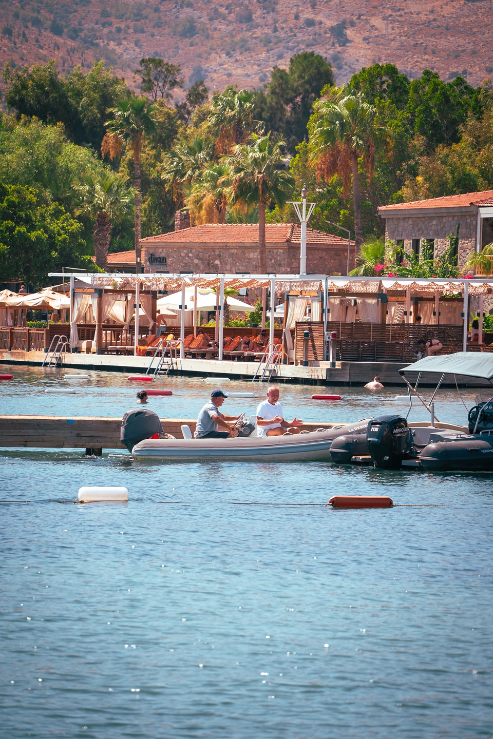 Two people in a grey dinghy by a jetty in Türkbükü, a beach club behind