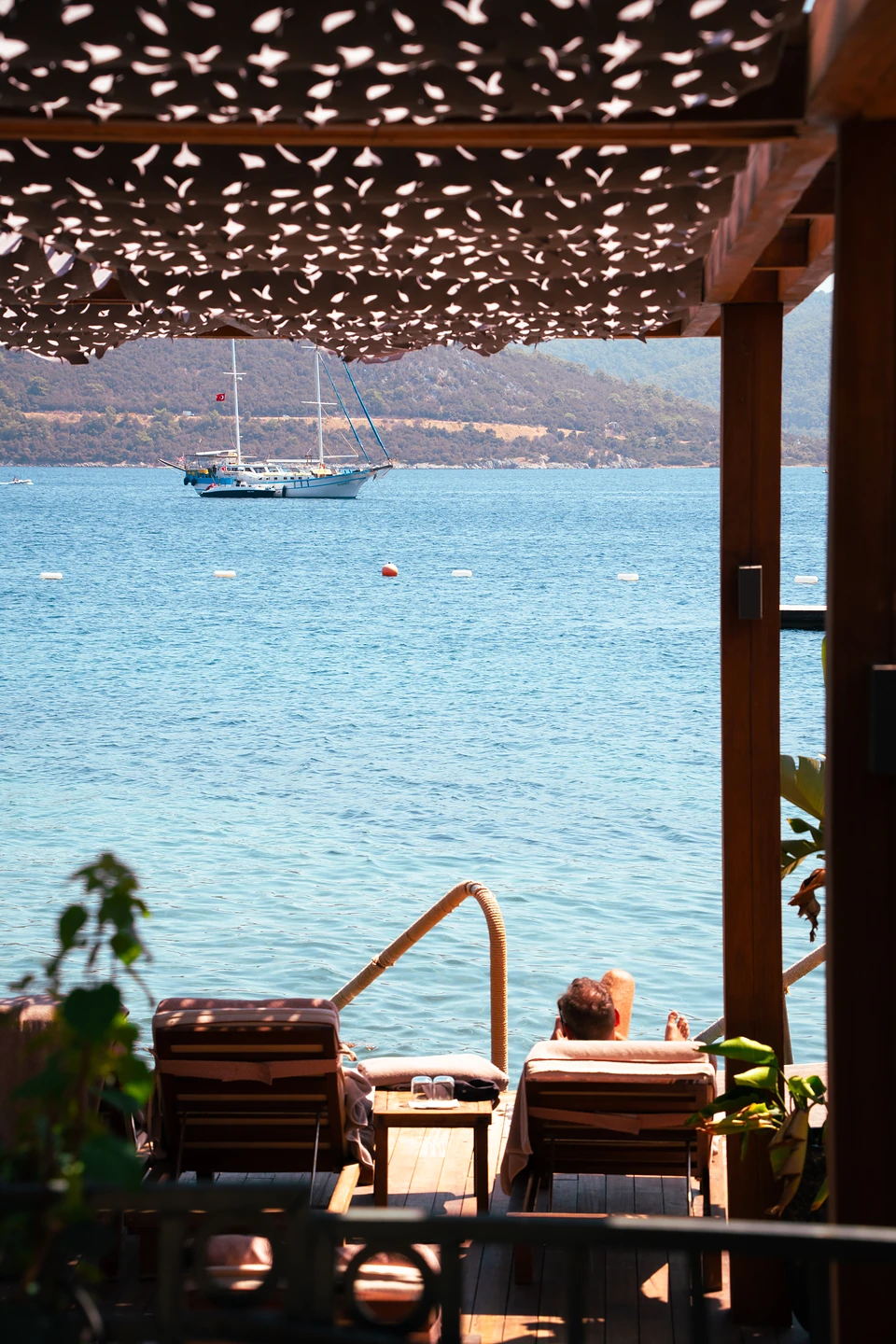 A person on a sunlounger on a shaded deck in Türkbükü, a gulet anchored offshore