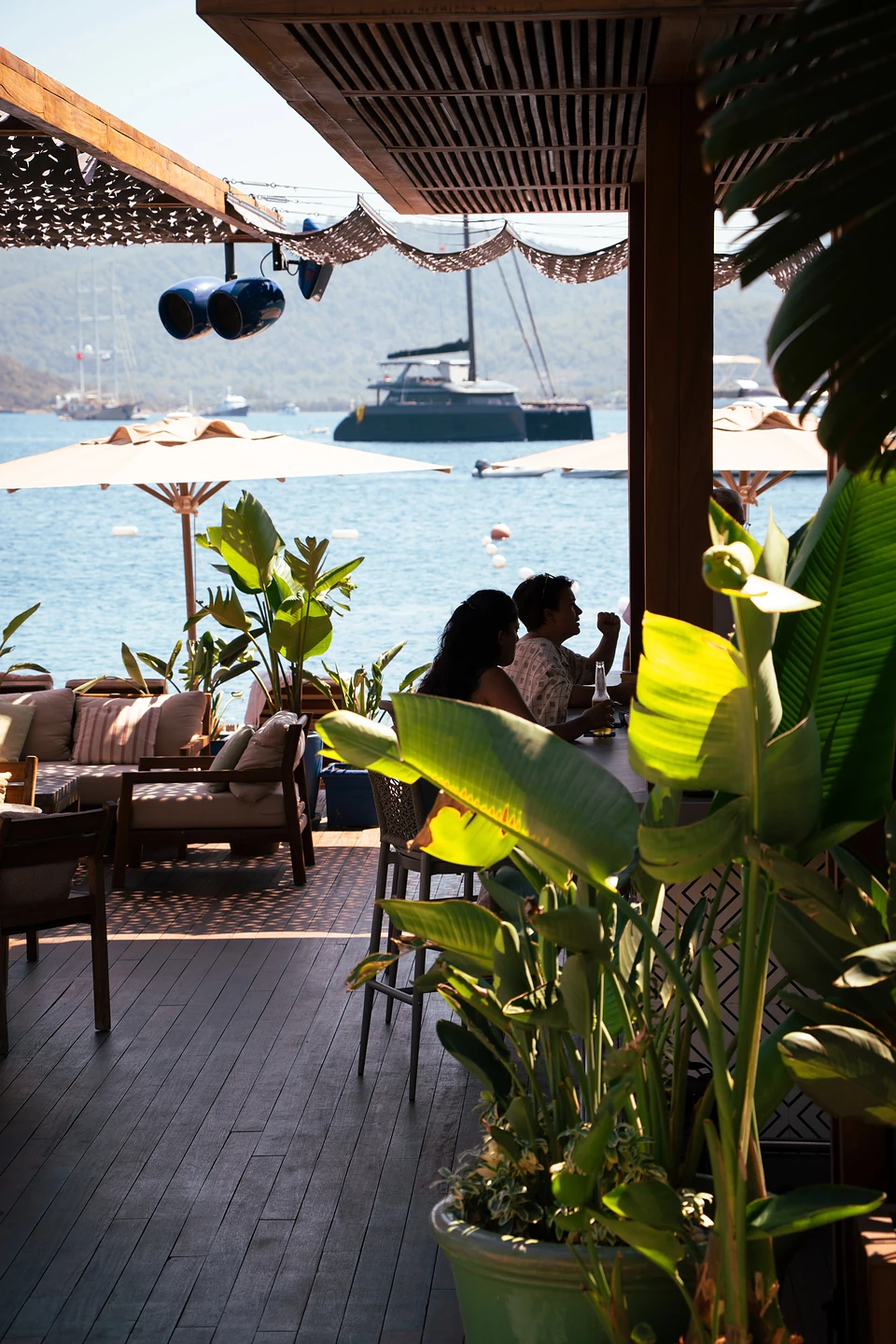 Two people at a shaded beach-club table in Türkbükü, yachts in the bay beyond