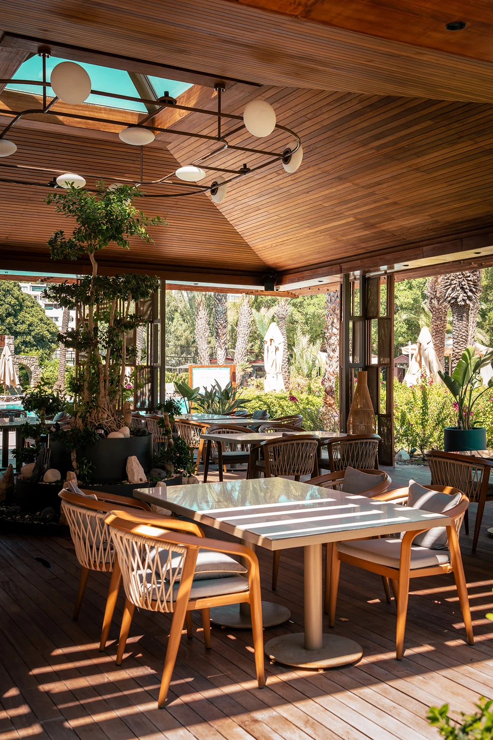 Rattan chairs at white tables under a wooden ceiling with globe lamps in Türkbükü, palms beyond