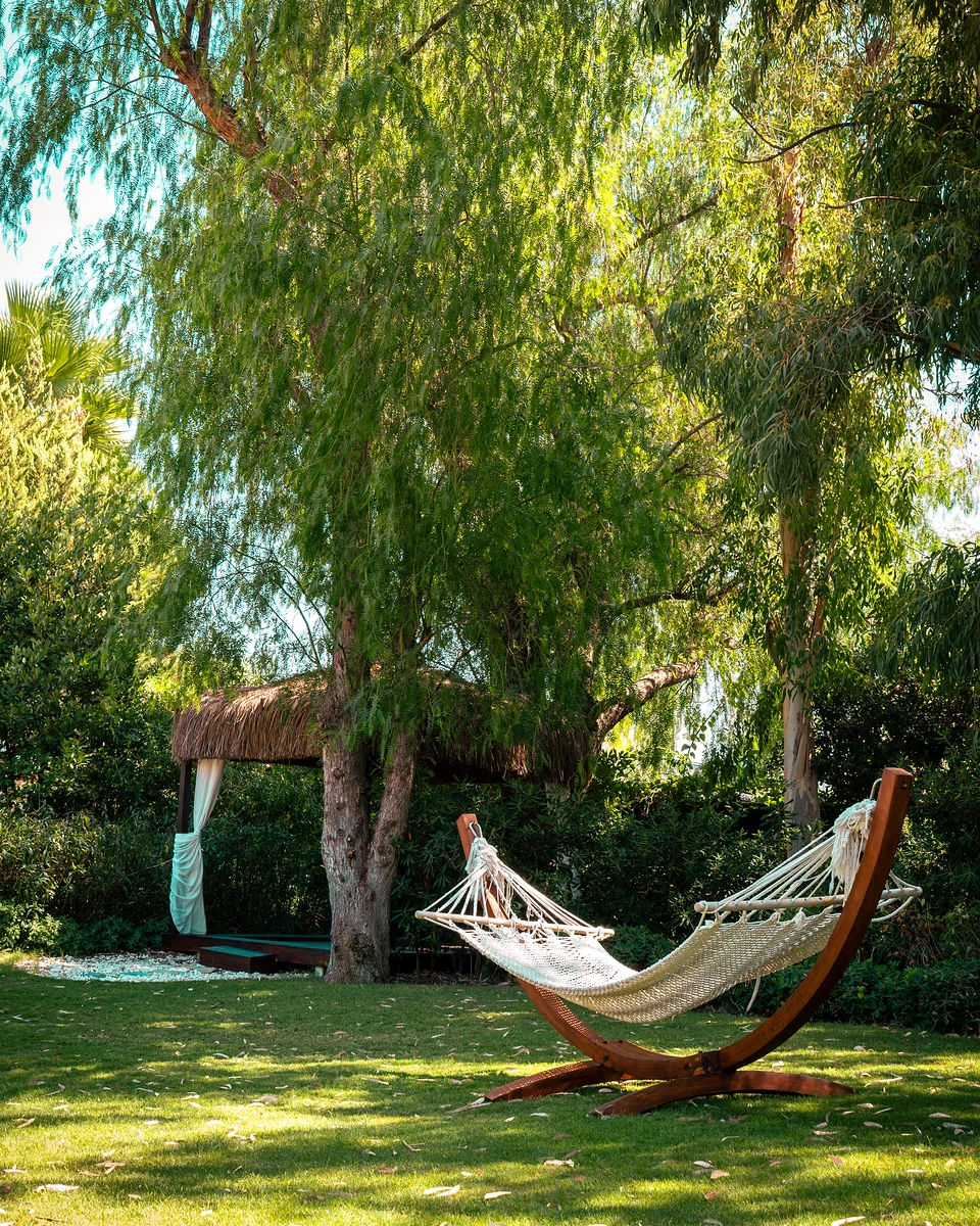 A rope hammock on a curved wooden stand on a Türkbükü lawn, a thatched cabana behind