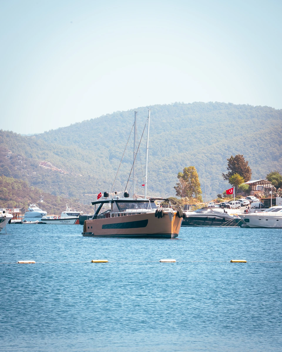 A tan-hulled two-masted yacht at anchor off Türkbükü, a Turkish flag on the shore
