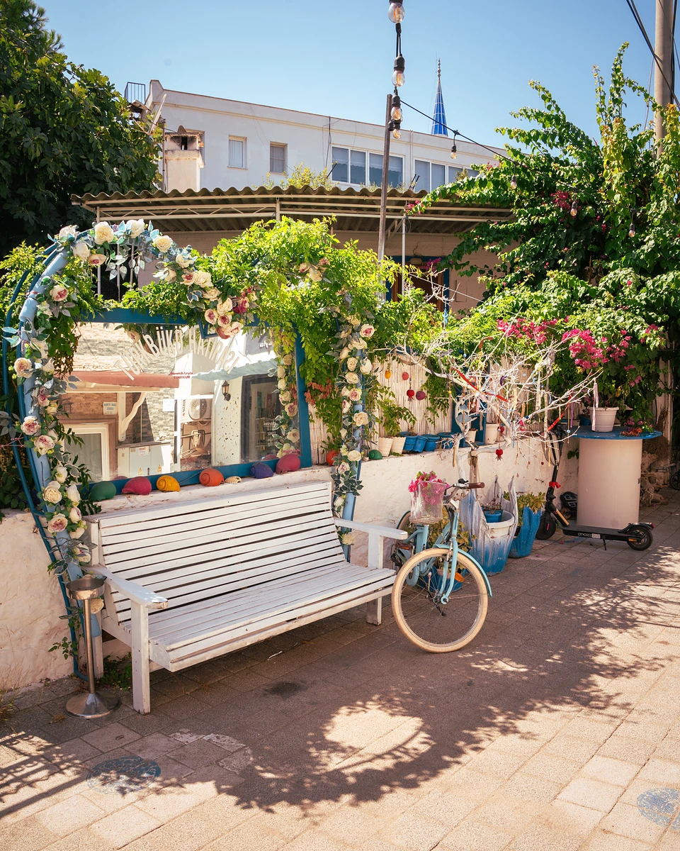 A white bench and a pale blue bicycle by a flower-garlanded mirror on a Türkbükü street