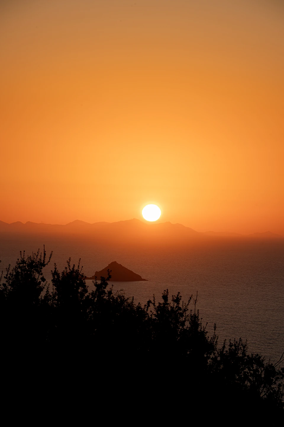 The sun sinking towards hazy mountains over the sea, a small pointed island in silhouette