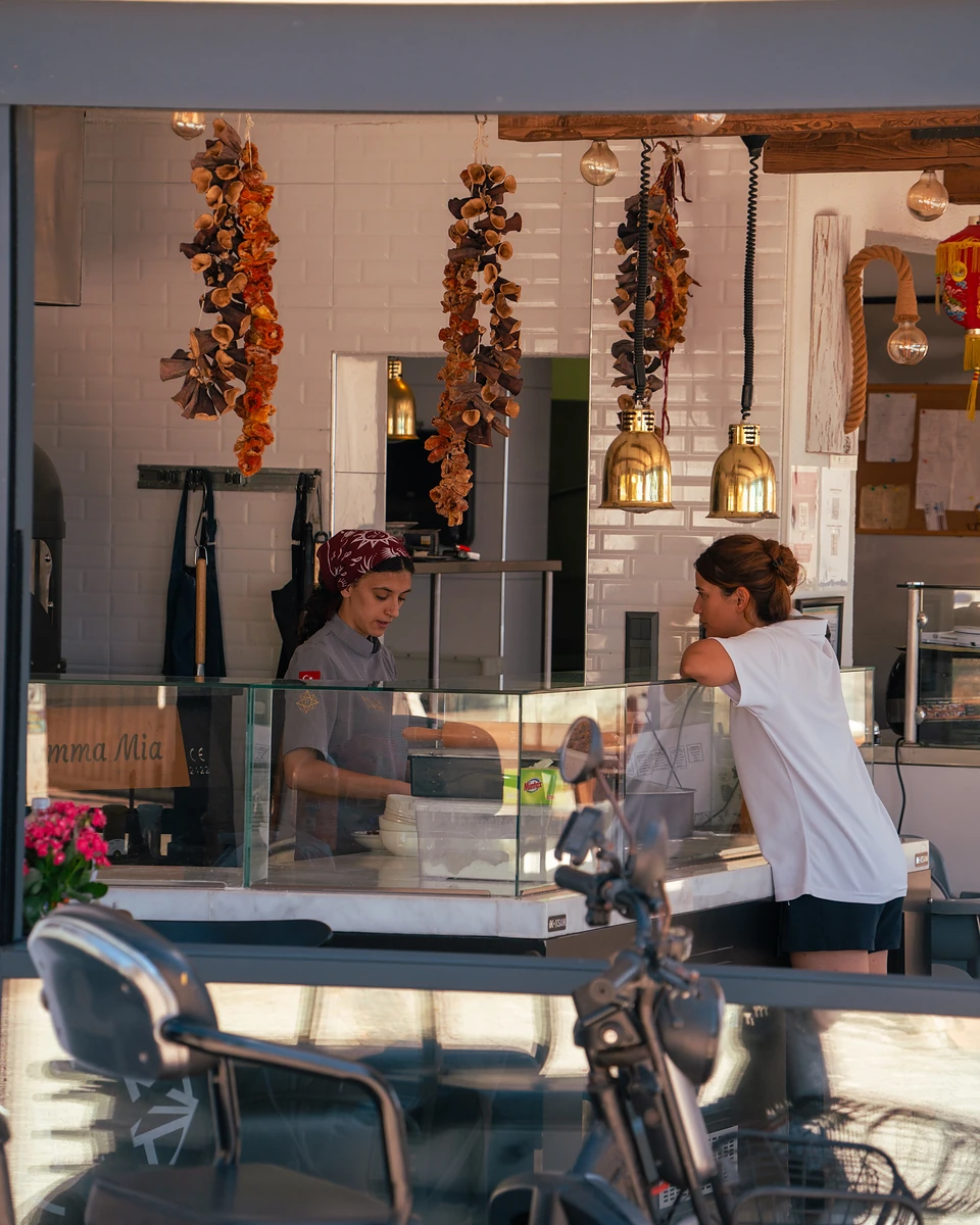 A cook in a red bandana at a glass counter in Yalıkavak, a customer leaning in