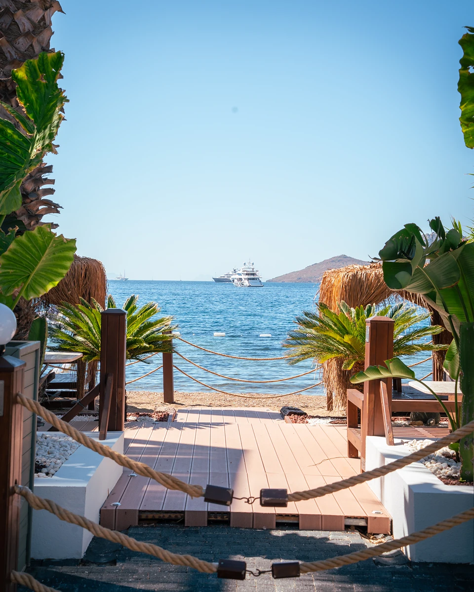 A boardwalk between rope rails leading to the beach in Yalıkavak, a yacht anchored offshore