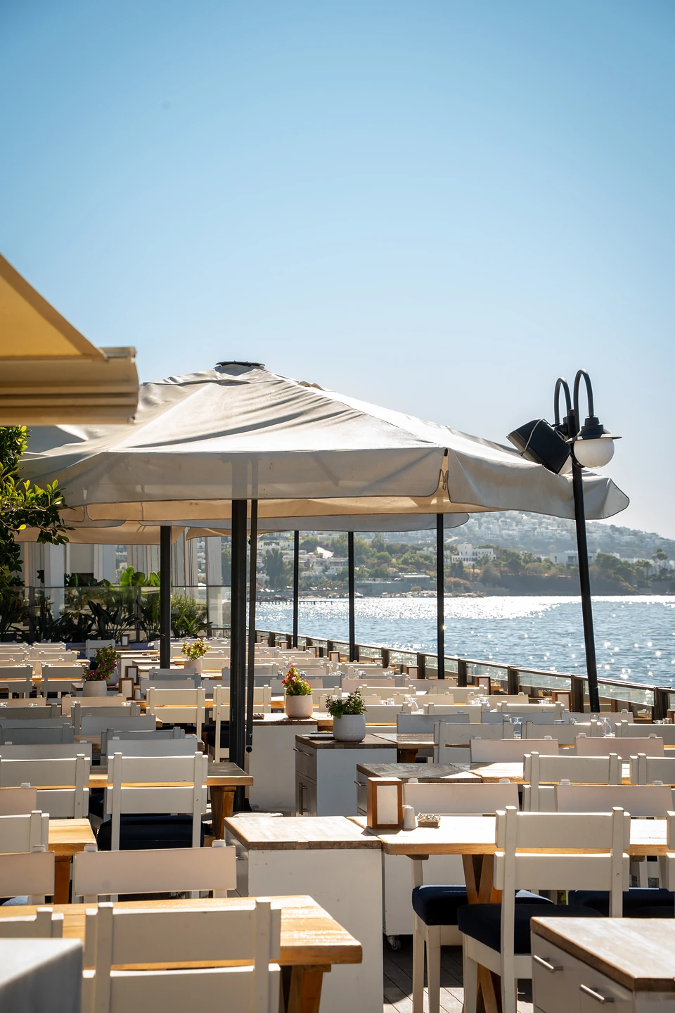 Rows of white chairs under big parasols on a seafront terrace in Yalıkavak, the sea glinting