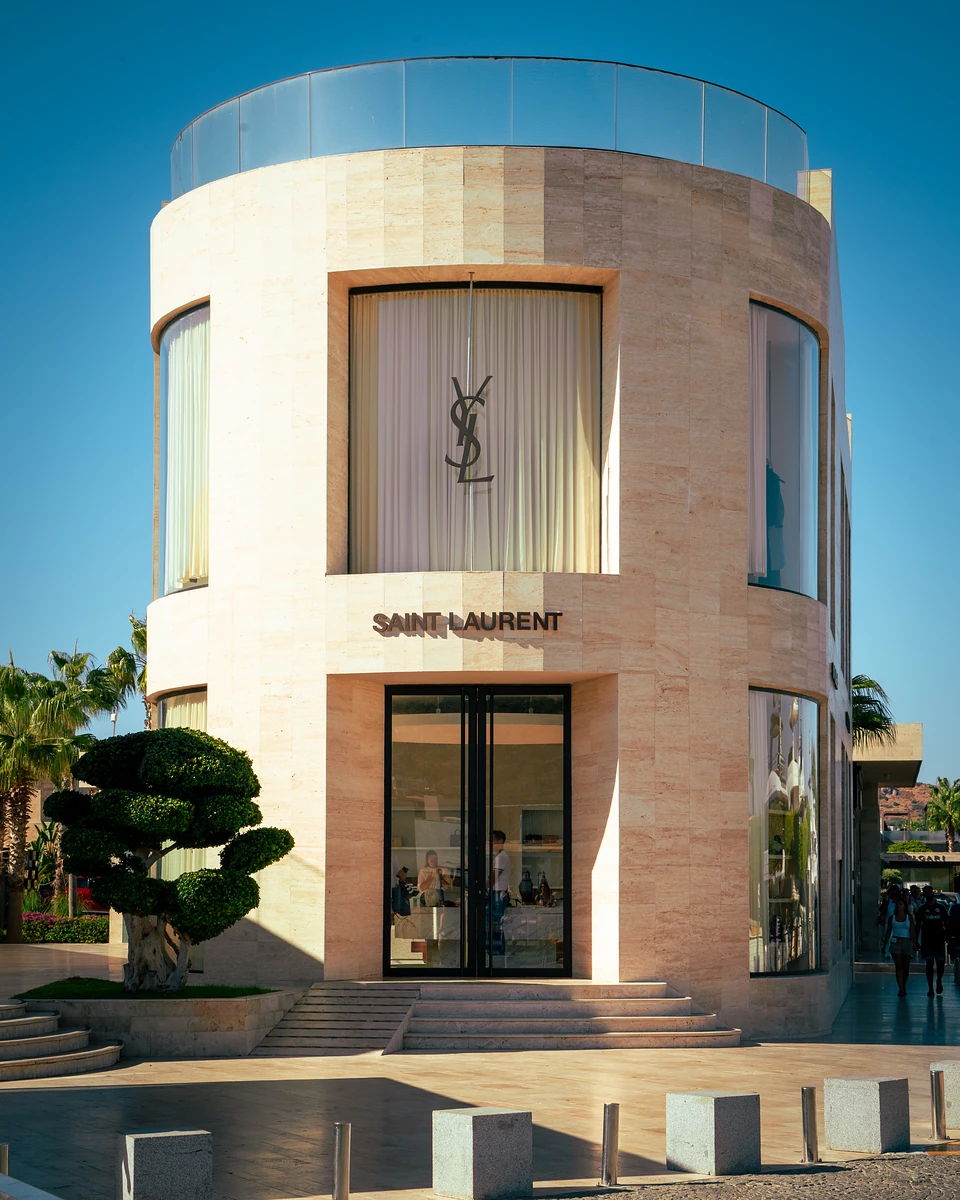 A curved stone shopfront with a lit sign in Yalıkavak, a clipped topiary tree beside