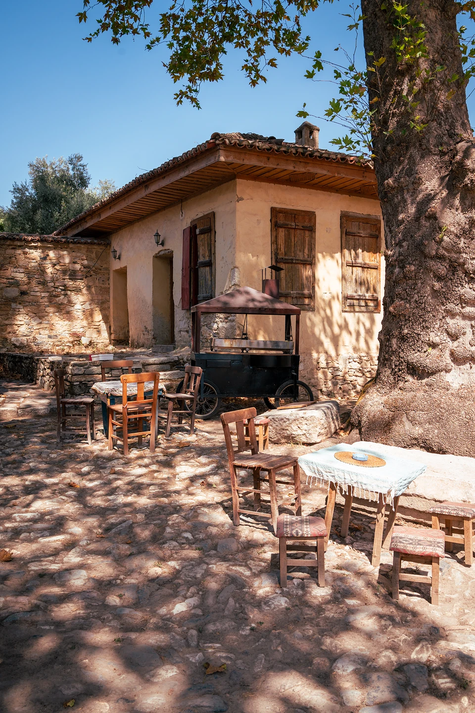 Wooden chairs and small tables in dappled tree shade before an old house with wooden shutters