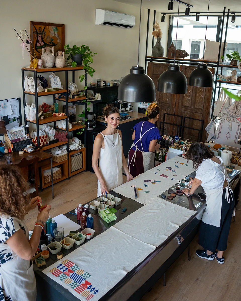 Four women in aprons around the long printing table, seen from above beside shelves of goods