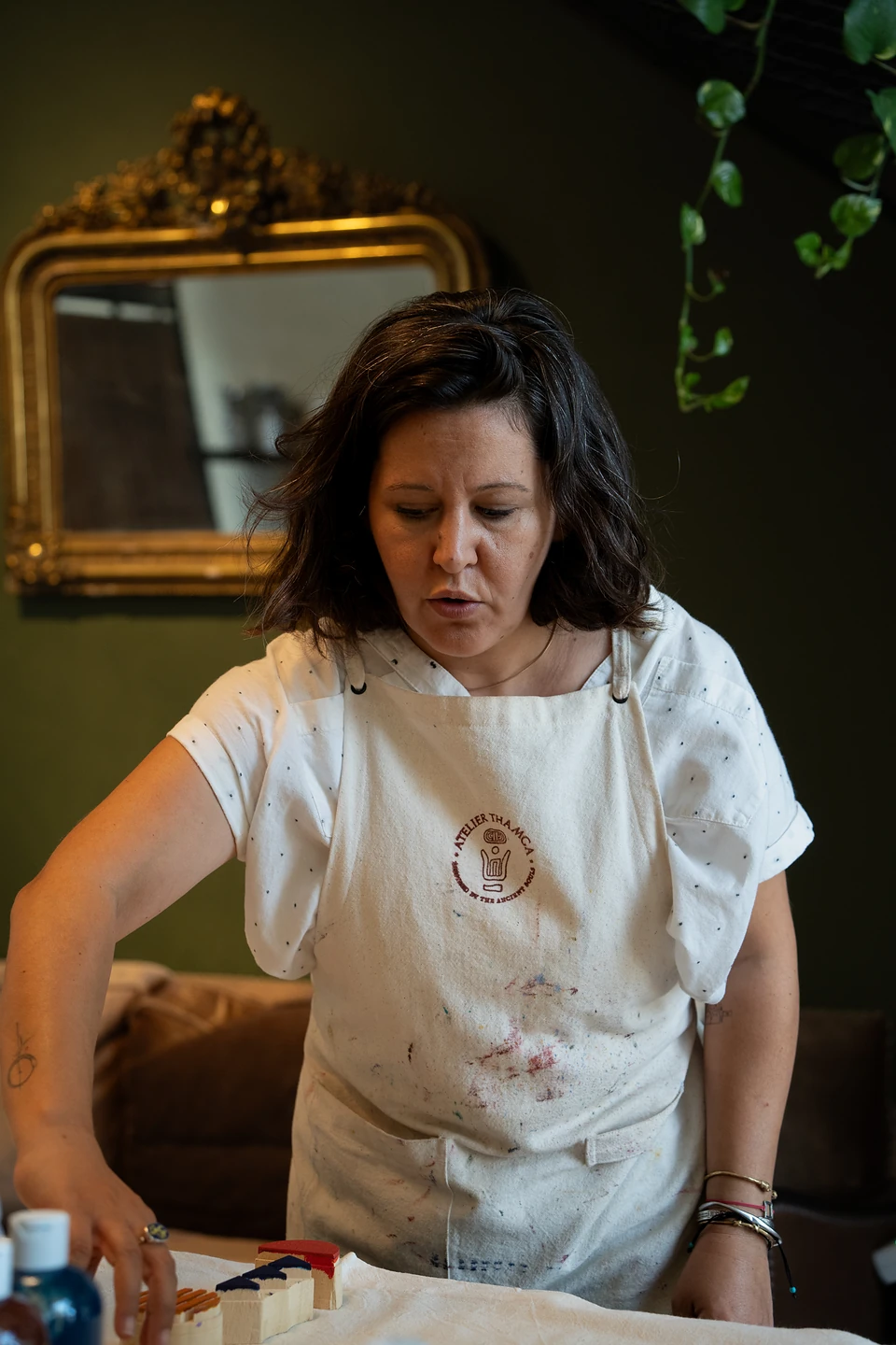 A woman in a paint-spattered Atelier Thamga apron reaching for blocks, a gilt mirror behind