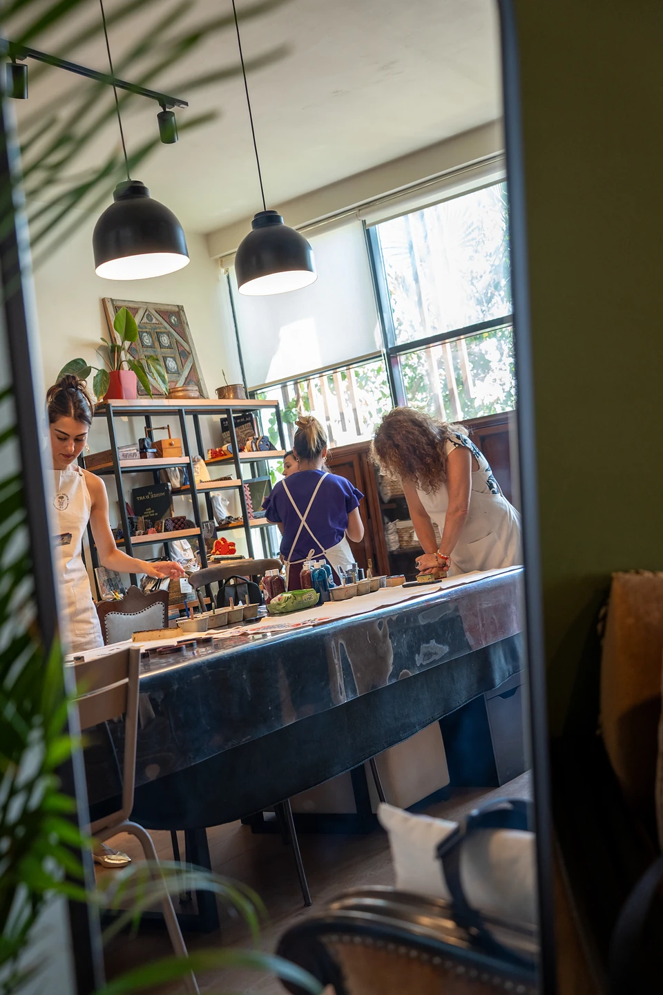 The workshop reflected in a tall mirror, three women at the long table under pendant lamps