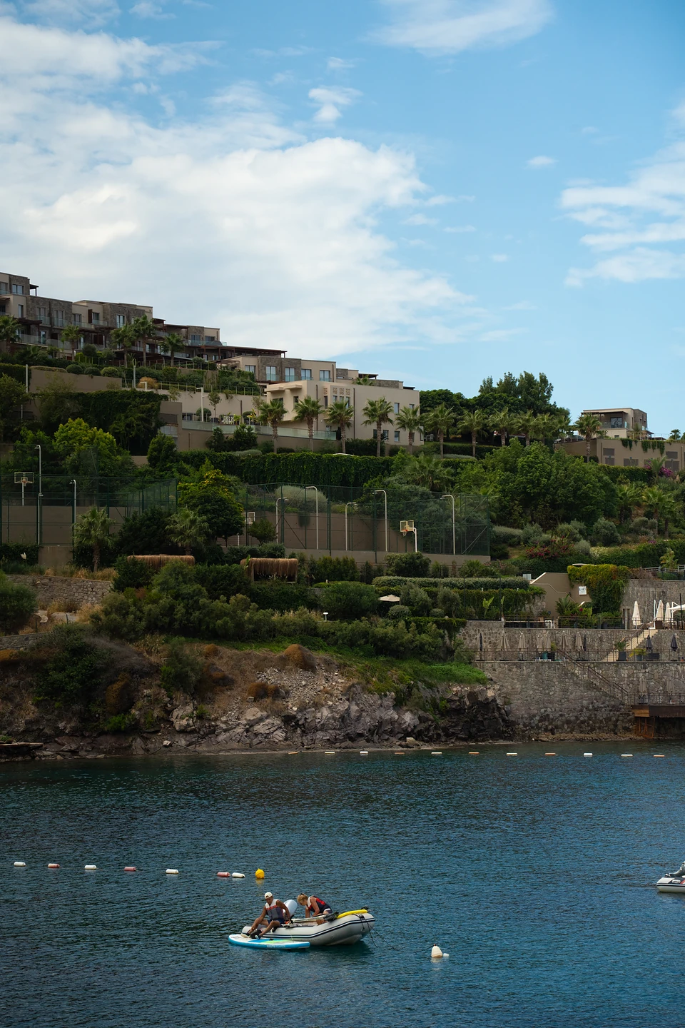 Two people in a dinghy in a calm Bodrum cove, terraced houses and palms above