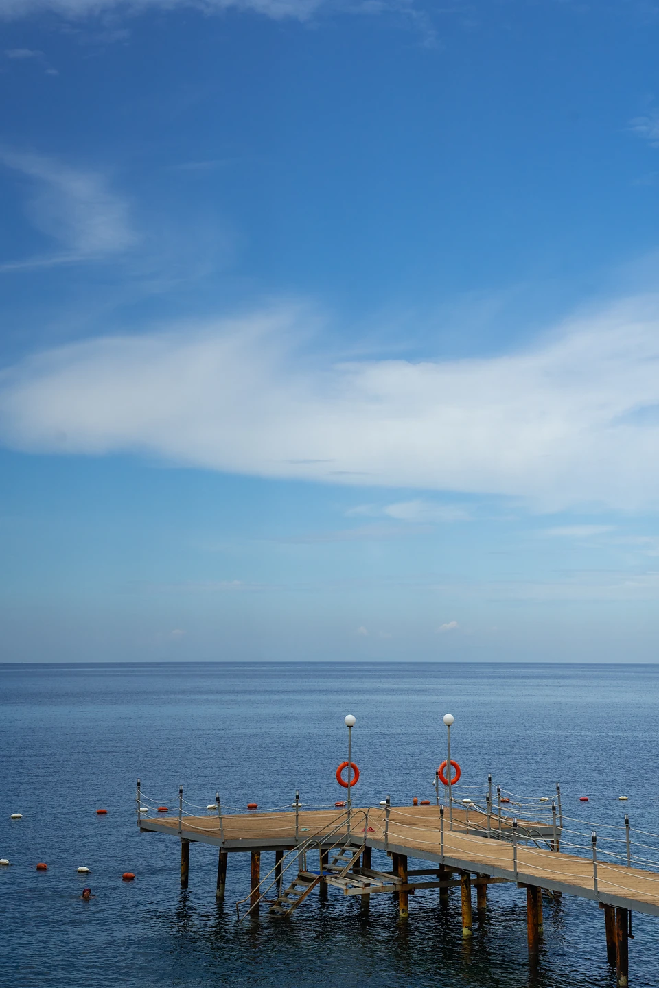 A wooden jetty on stilts with two orange lifebuoys, reaching into a flat calm sea
