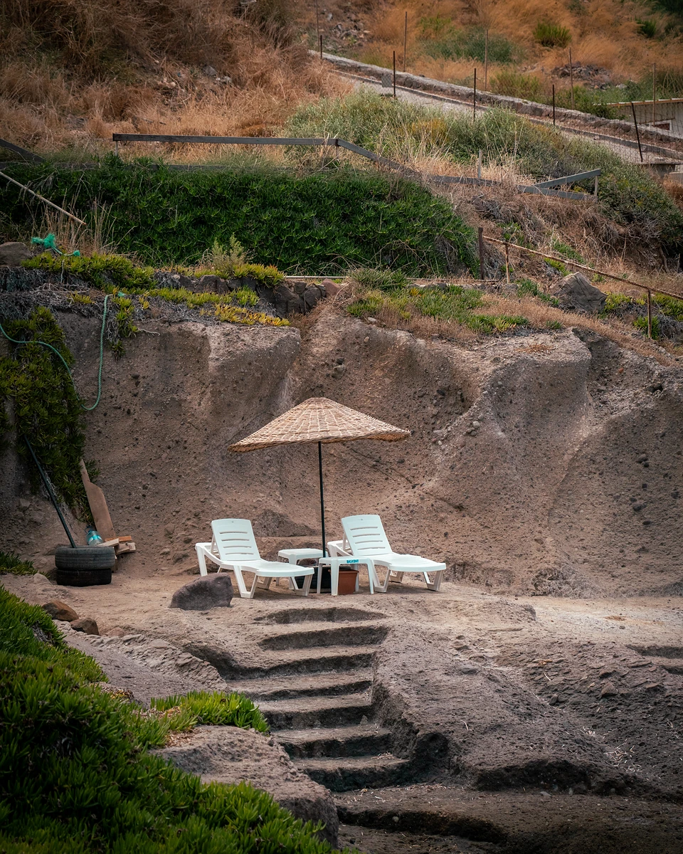 Two white sunloungers under a woven parasol on a sandy ledge, rough steps below