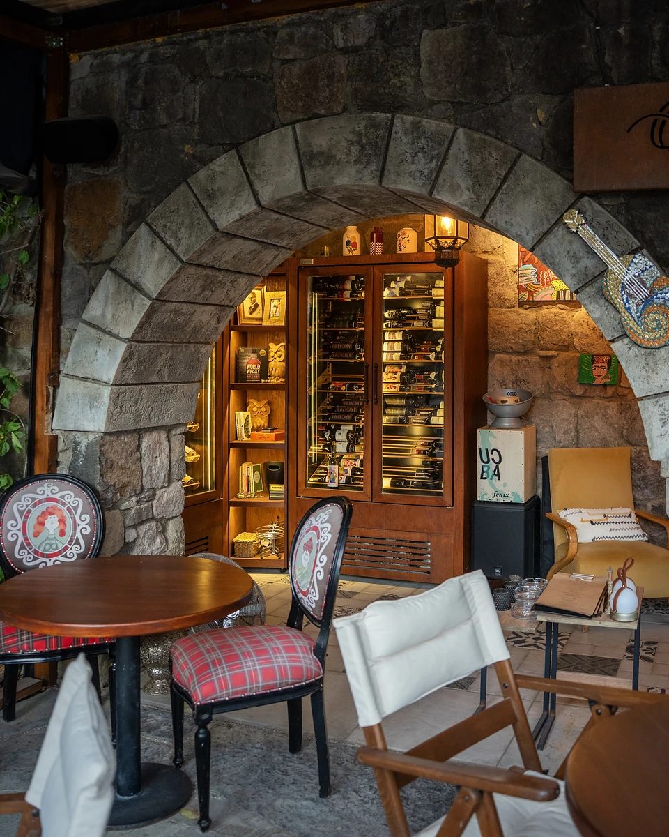 A wine cabinet lit beneath a stone arch, a tartan-cushioned chair and round table in front
