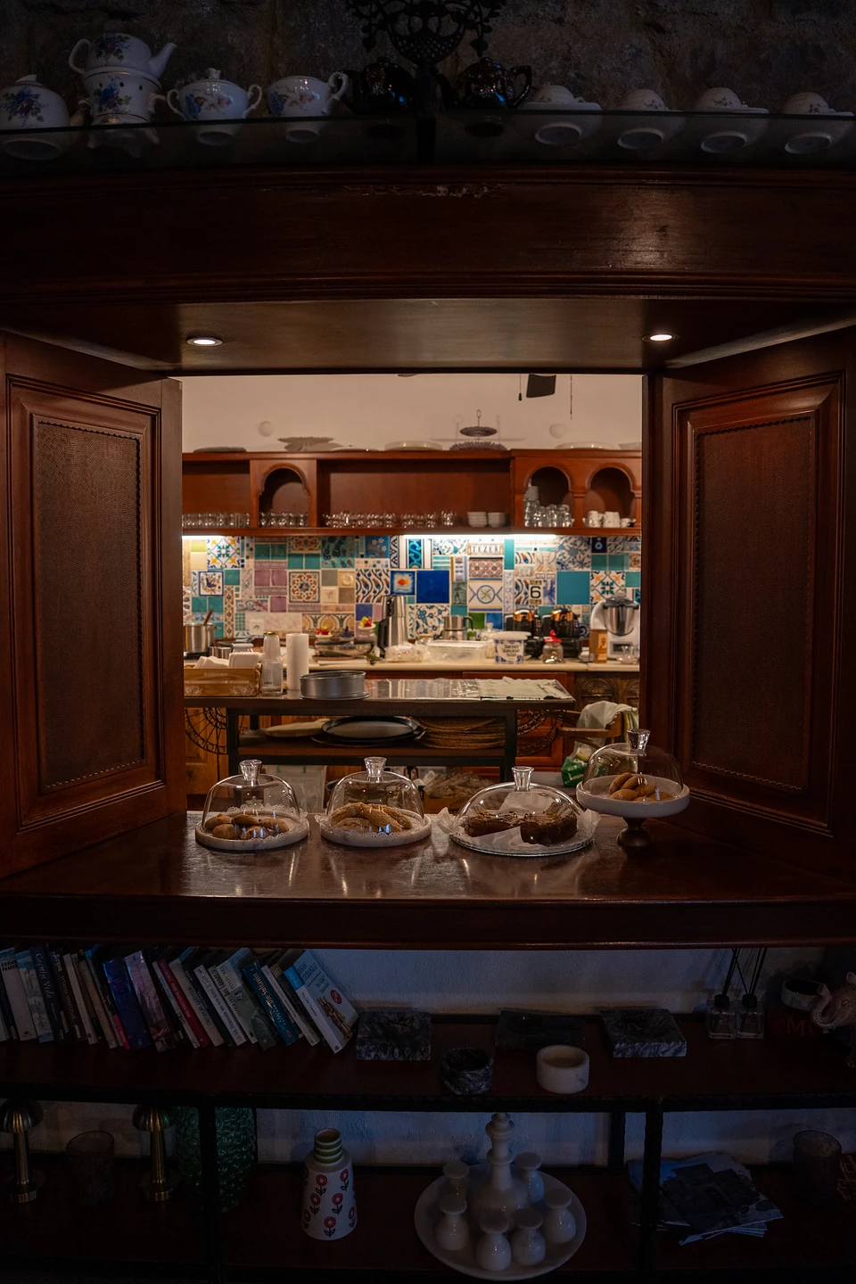 Biscuits and cake under glass domes in a serving hatch, a kitchen of colourful tiles beyond