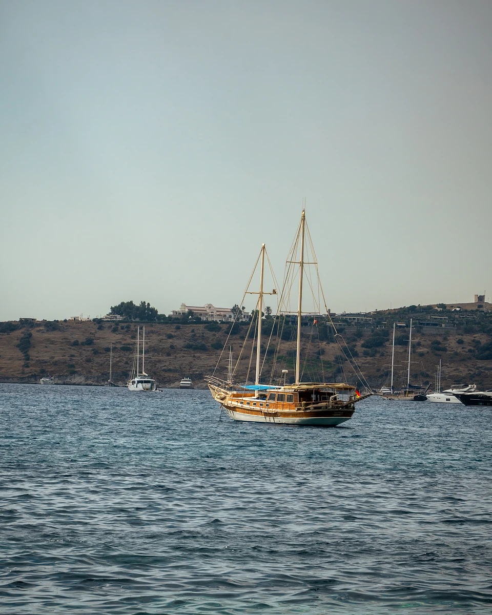 A wooden two-masted gulet at anchor off Bodrum, a dry hillside dotted with villas behind