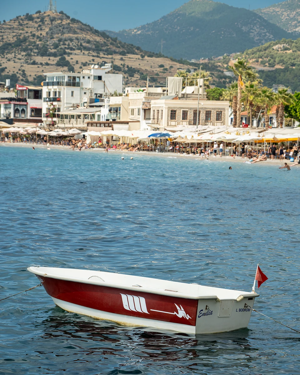 A red and white boat named ‘Emilia’ moored off a busy town beach in Bodrum