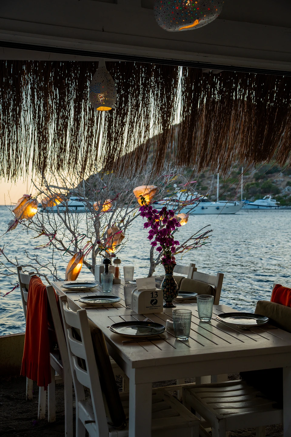 A table laid at the water’s edge in Gümüşlük at dusk, purple flowers and lanterns