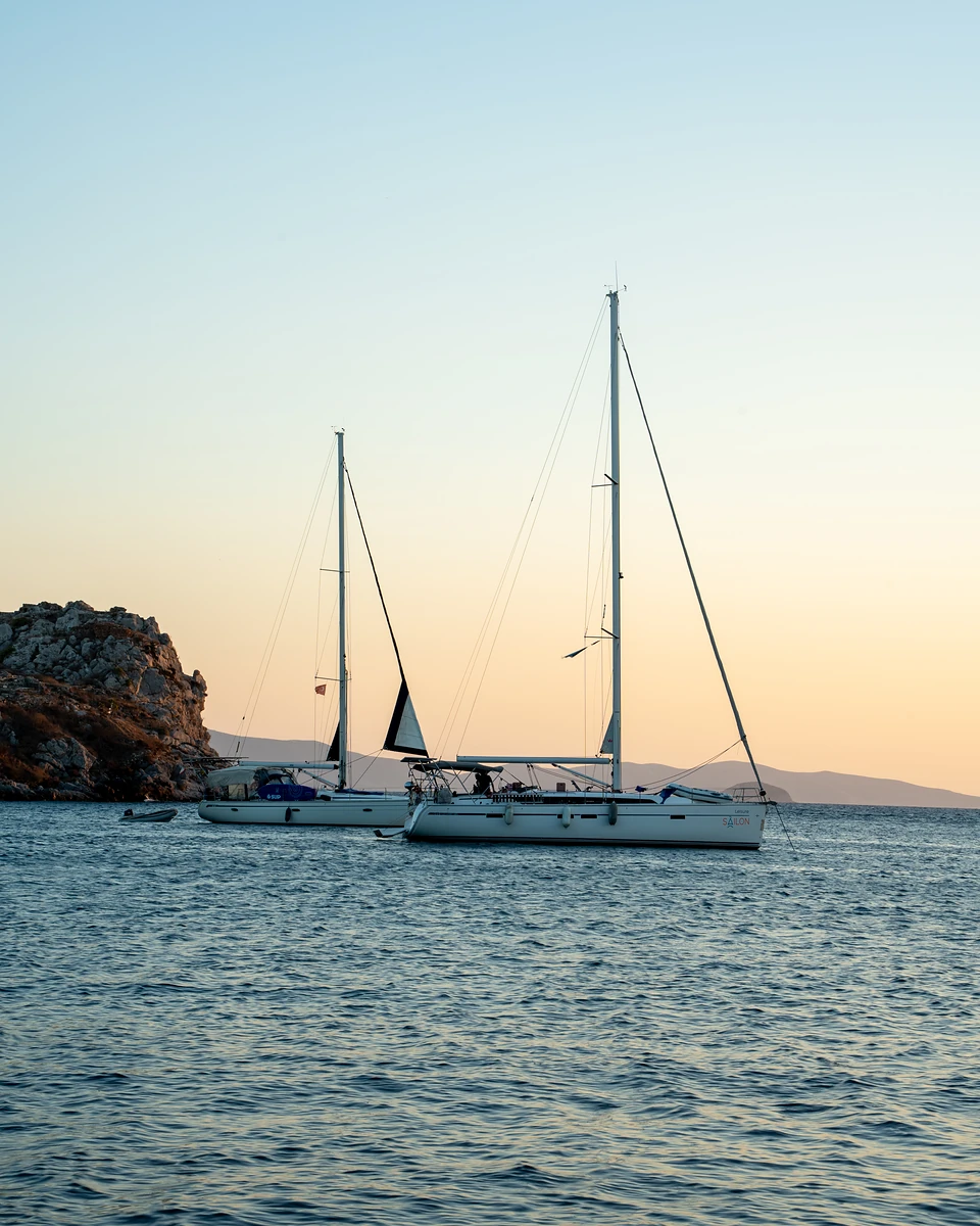Two sailing yachts at anchor off a rocky headland in Gümüşlük, the dusk sky turning peach