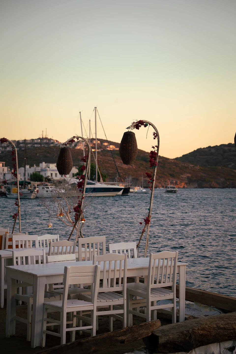 White wooden tables at the water’s edge in Gümüşlük, woven lanterns on poles at sunset