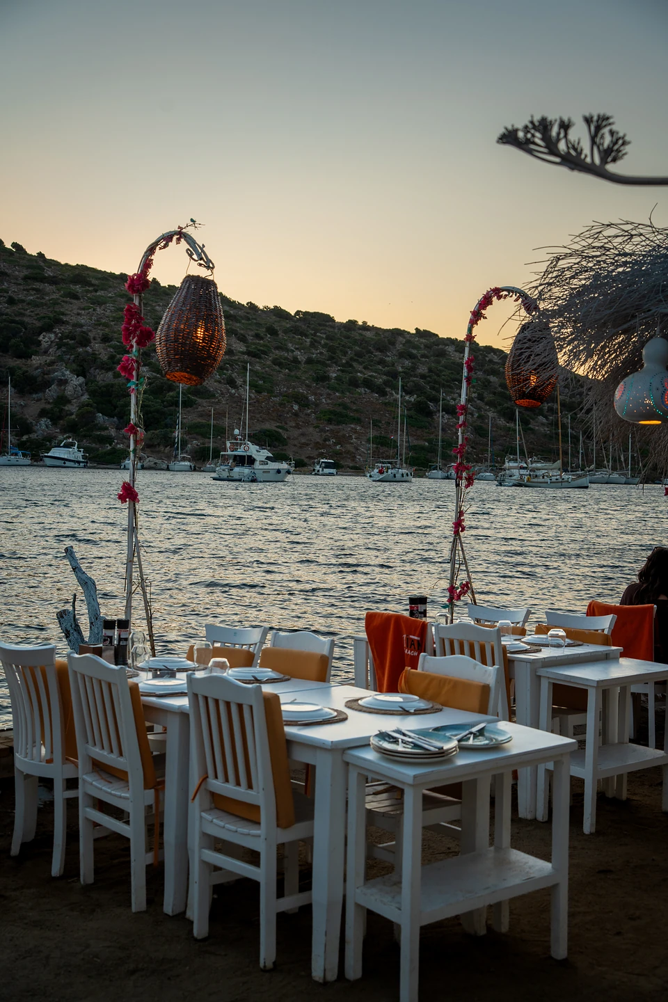 Tables with orange cushions and lit woven lanterns by the bay in Gümüşlük, yachts beyond