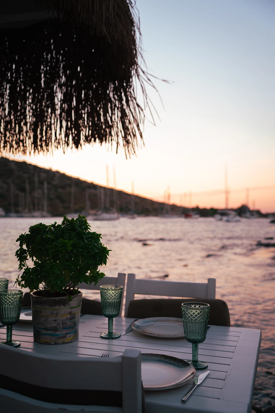 Green cut-glass goblets and a potted plant on a waterside table in Gümüşlük at dusk