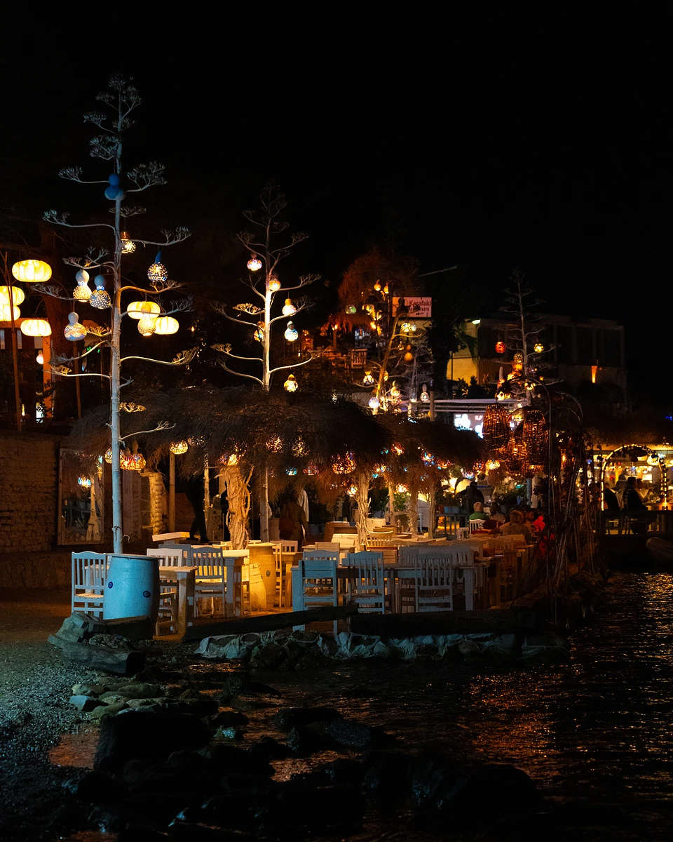 Gümüşlük’s waterfront at night, white tables under gourd lamps hung from tall dried stalks