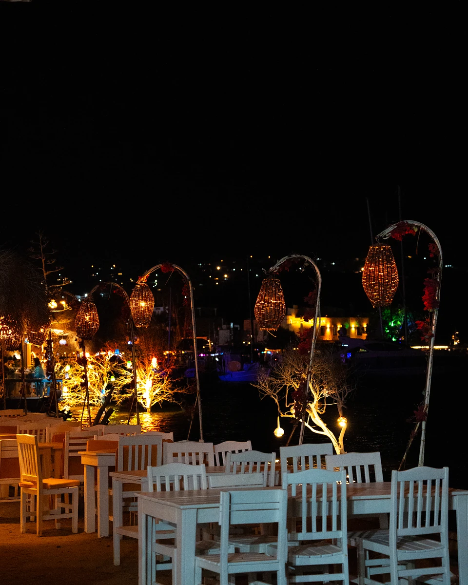Empty white tables by the water in Gümüşlük at night, woven lanterns glowing on curved poles