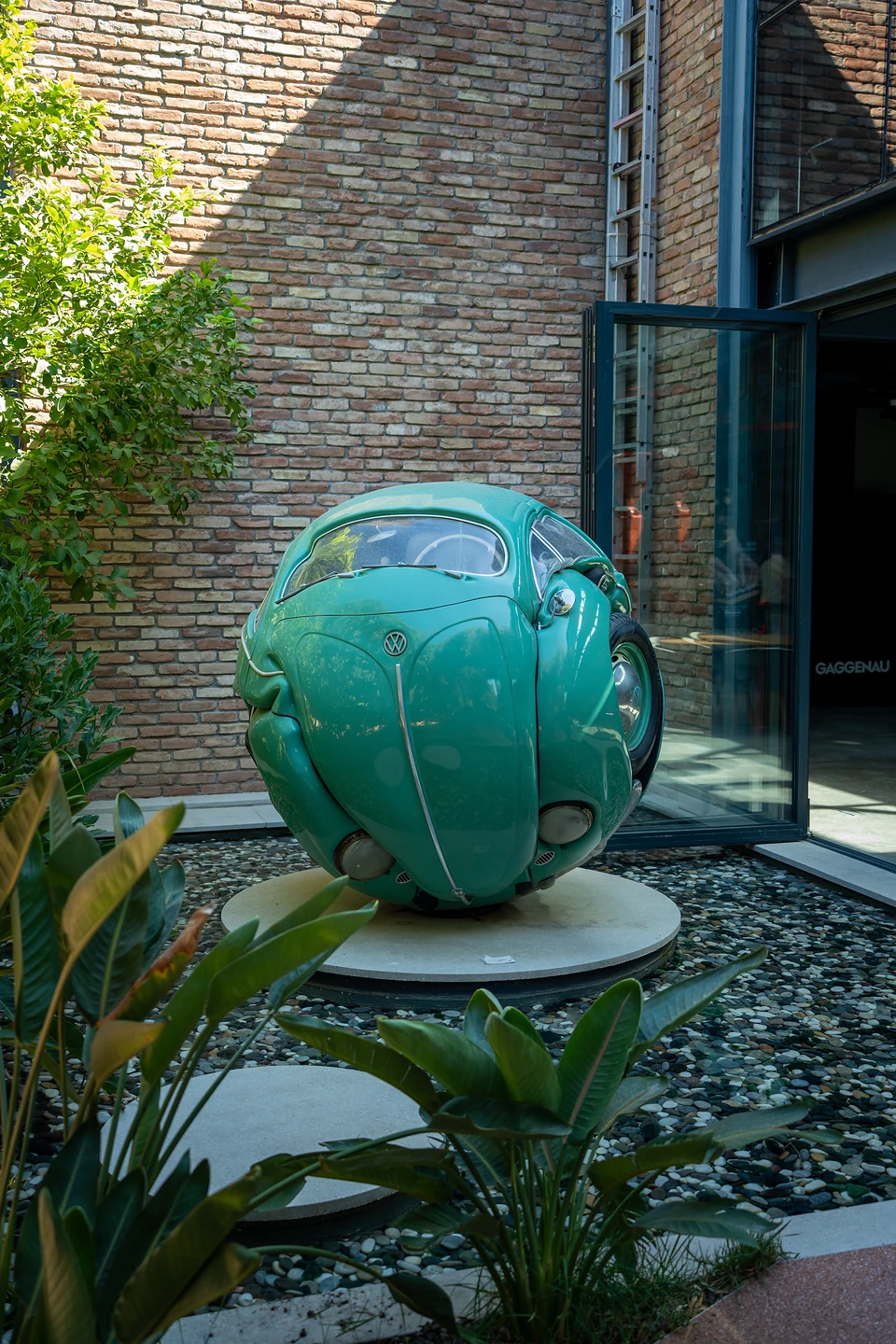 A green Volkswagen Beetle sculpted into a ball, on a pebble bed by a brick wall