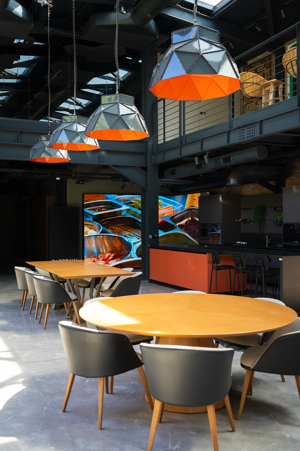 Faceted lamps with orange undersides over wooden tables in the glass hall, a kitchen beyond