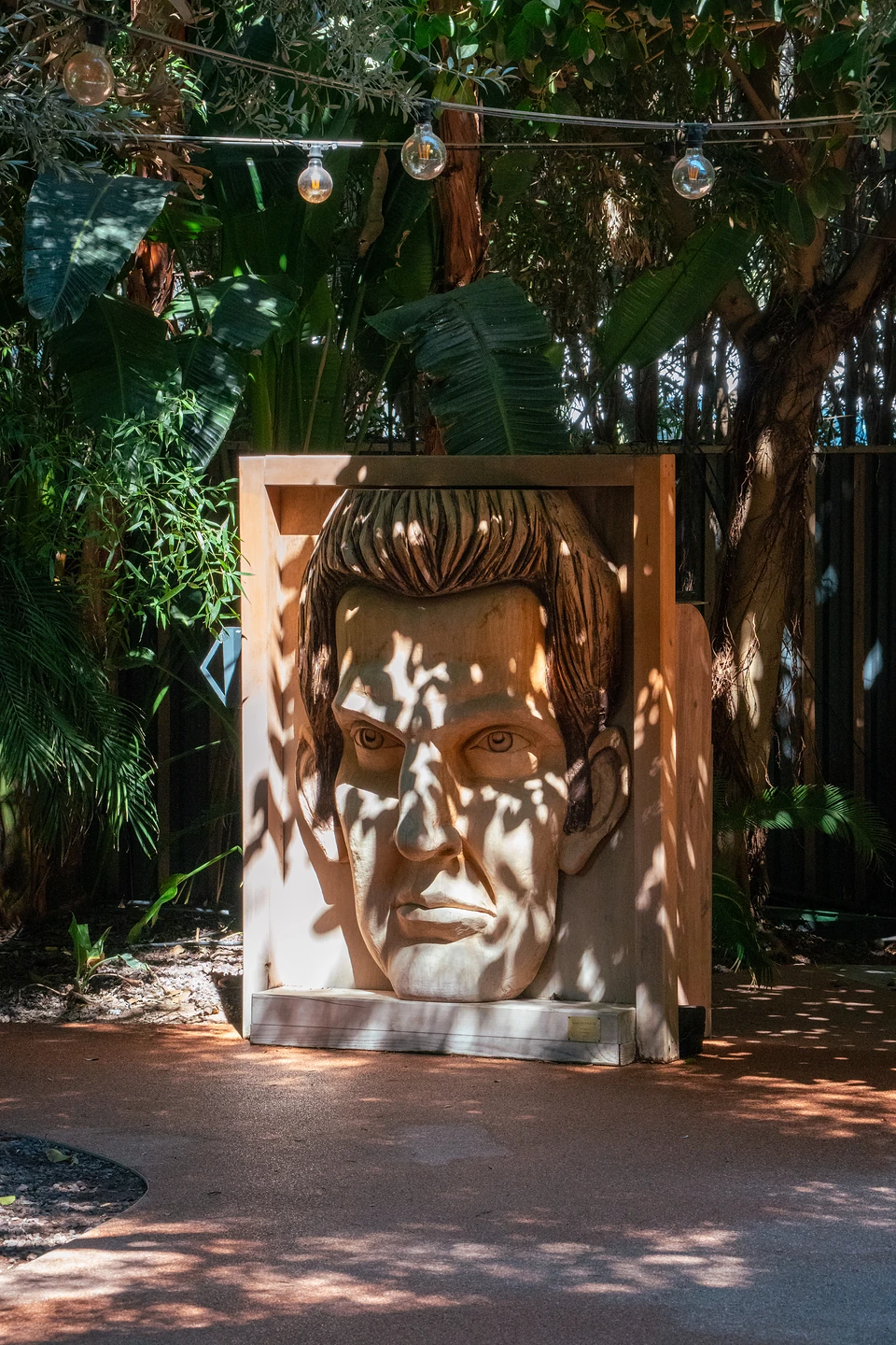 A giant carved wooden face in a box frame, dappled with leaf shadows under string lights