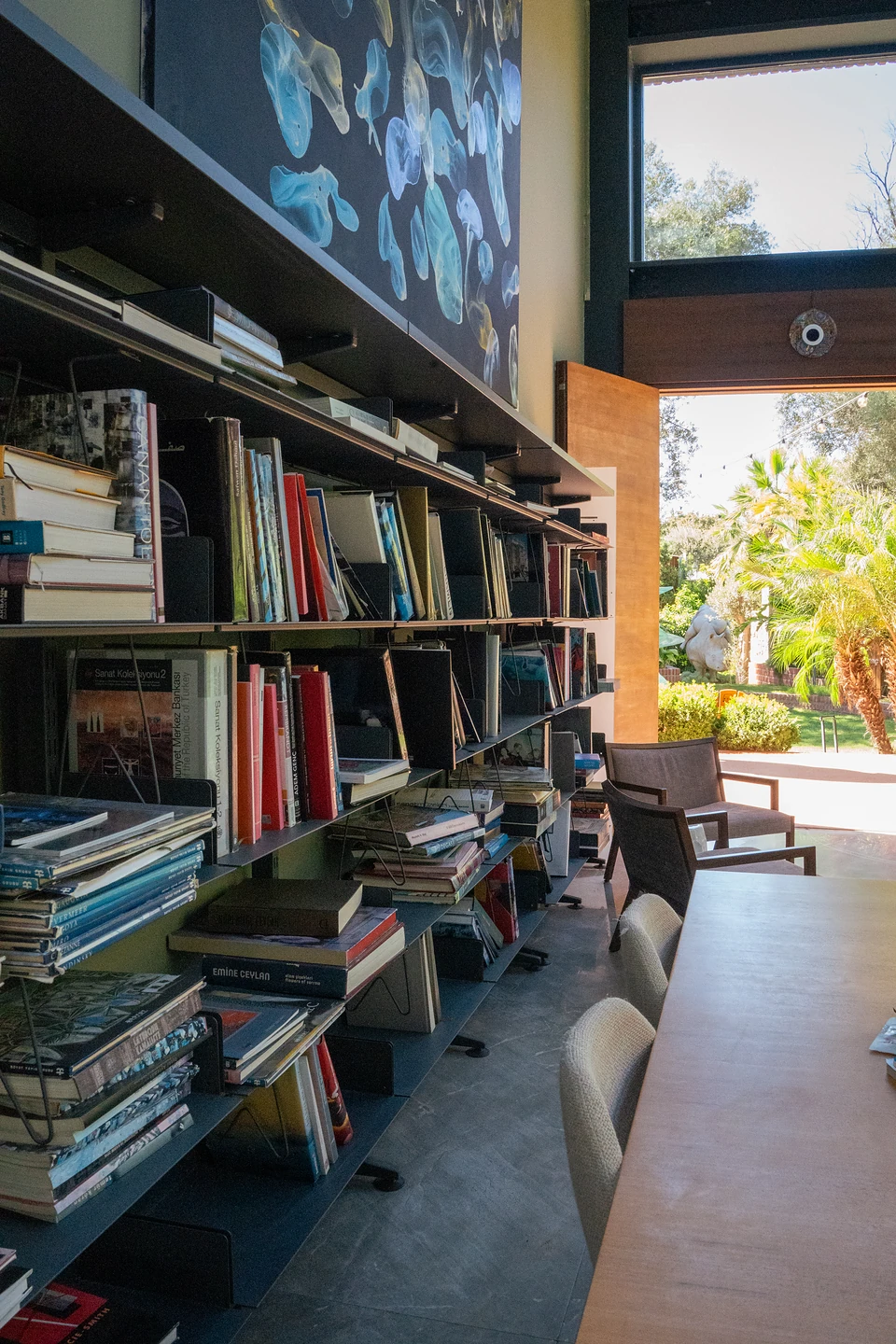 Packed bookshelves beside a long table in Zai's library, a door open to the sunlit garden