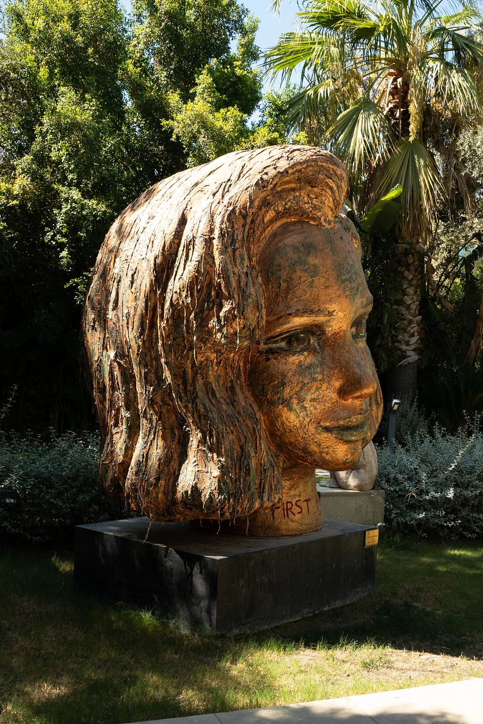 A giant copper-toned head of a woman with flowing hair on a black plinth, palms behind