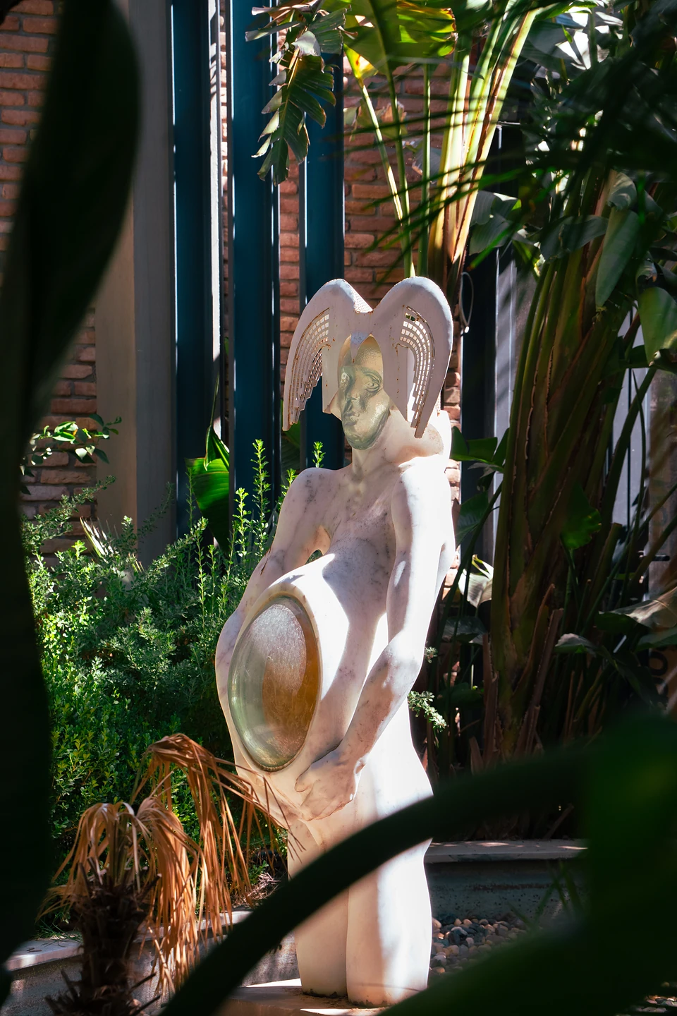 A white sculpted figure with a gilded face, winged headdress and golden disc, seen between leaves