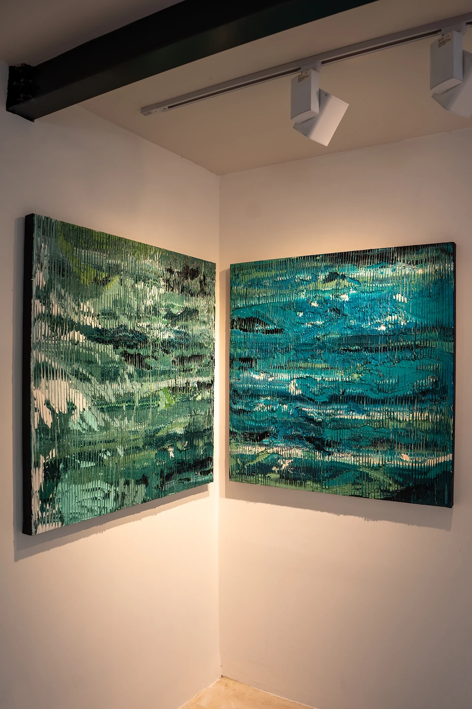 Two textured paintings in greens and teal, like rippling water, hung in a white gallery corner