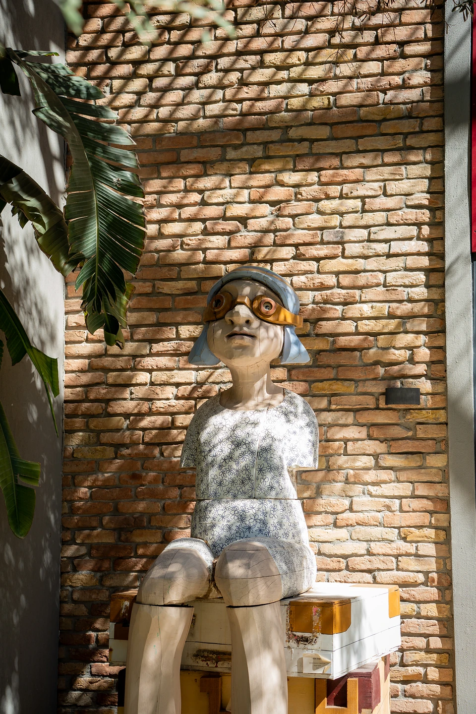 A carved wooden figure in copper goggles seated on painted boxes against a sunlit brick wall