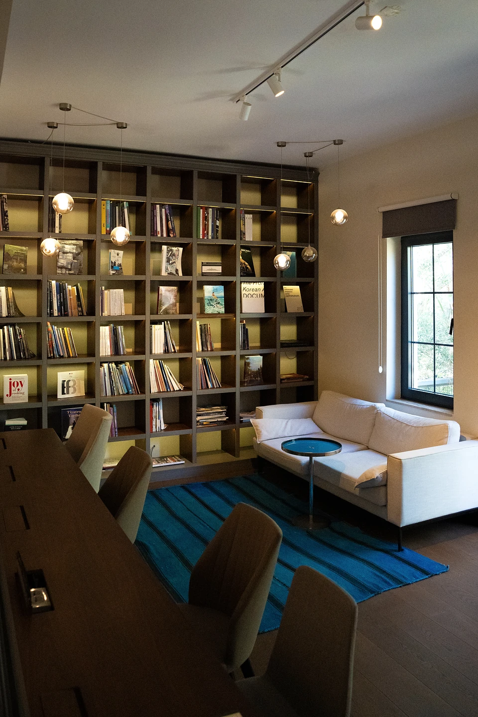 A reading room: a tall bookcase, a cream sofa on a blue rug, glowing globe lamps