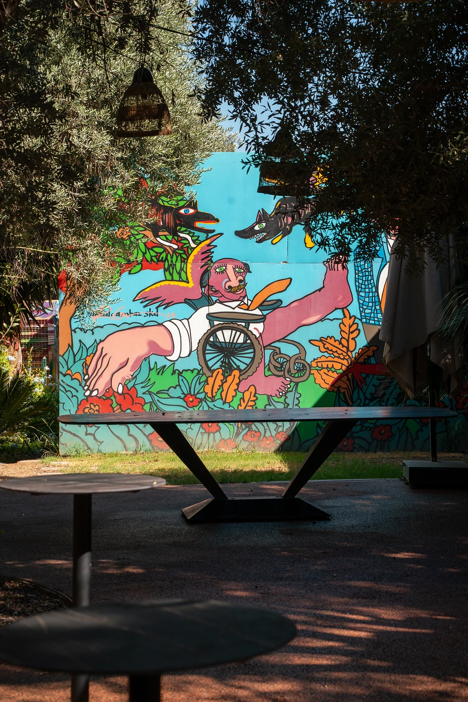 A bright mural of a winged figure, a wheel and flowers, seen past an empty table