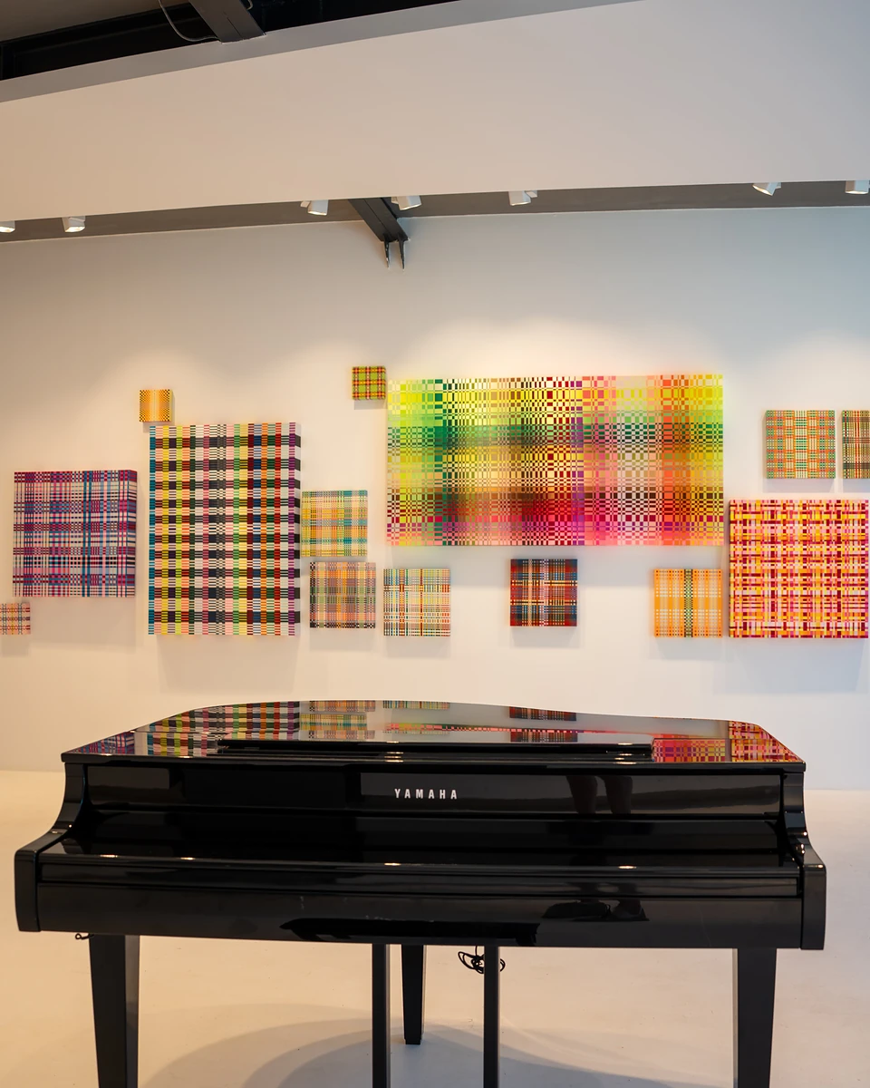 A black grand piano before a white wall hung with plaid canvases in bright colours