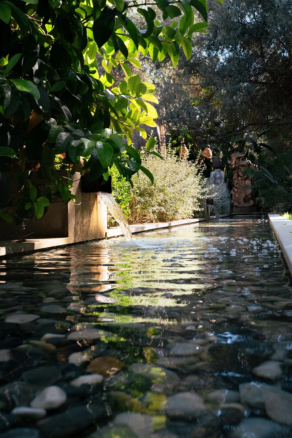 A pebble-bottomed water channel in the garden, sunlit leaves above and statues at the far end
