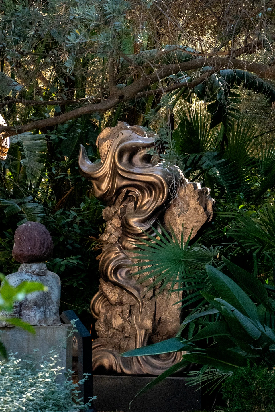 A swirling polished metal form wrapped around rough rock, framed by palms and olive branches