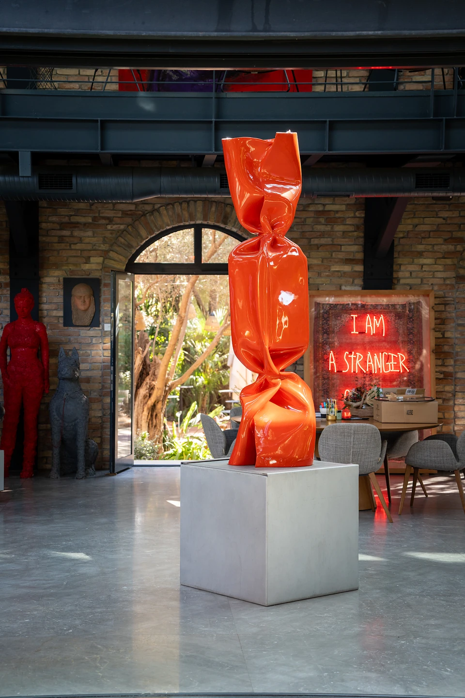 A tall red wrapped-sweet sculpture, with a neon reading “I am a stranger” glowing behind
