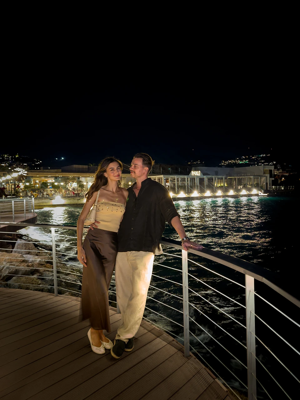 The two of us on a seafront boardwalk at night in Yalıkavak, lights on the water