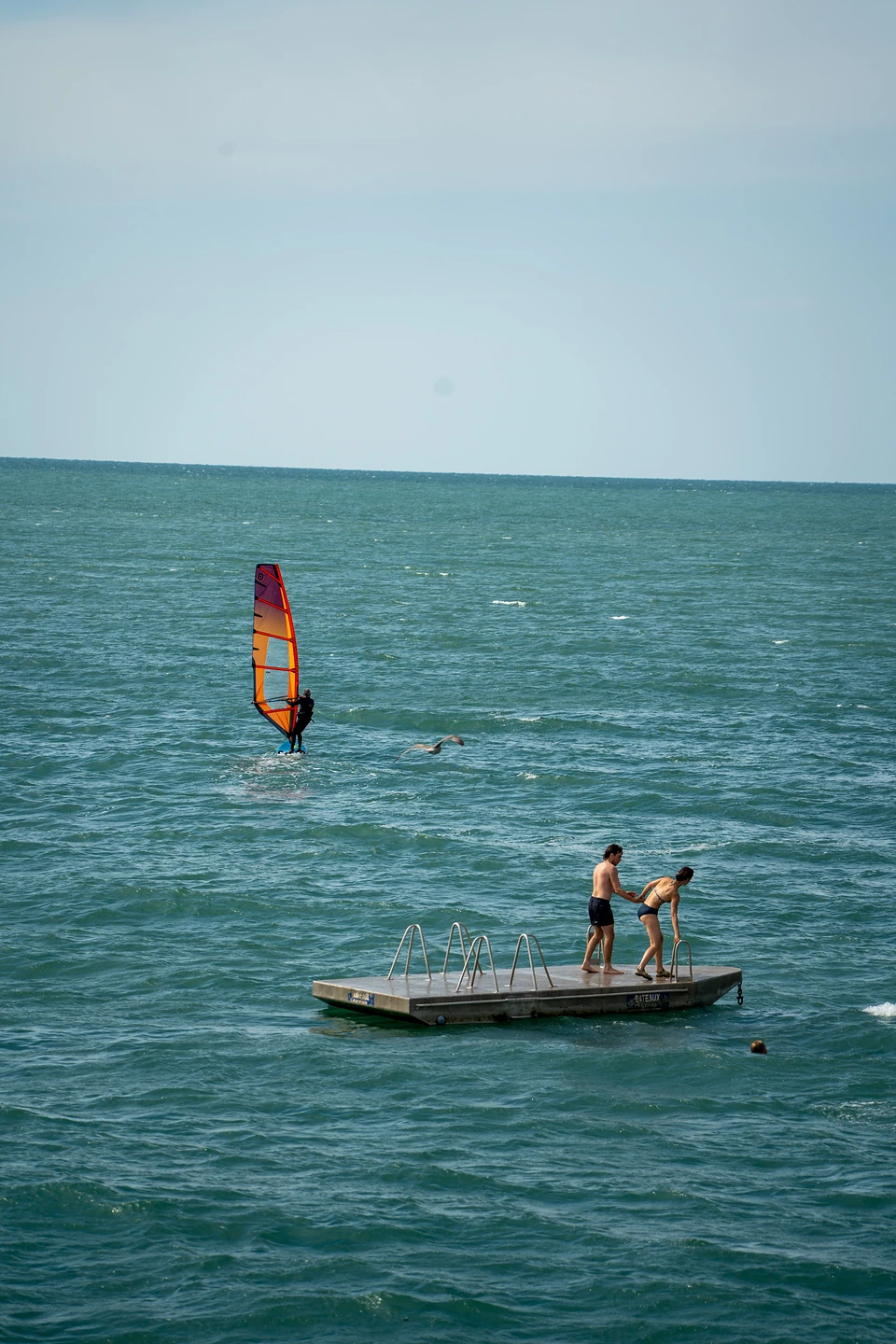 A windsurfer with an orange sail beyond two people on a floating swim platform