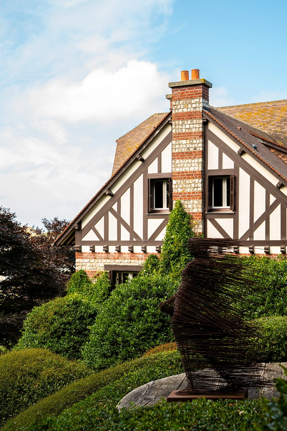 A half-timbered house behind clipped hedges and a woven metal sculpture at Les Jardins d'Étretat