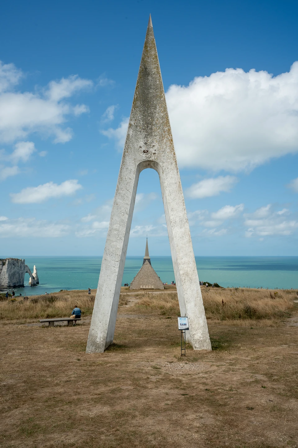A tall pointed white monument framing the clifftop chapel, the arch and the Needle far left