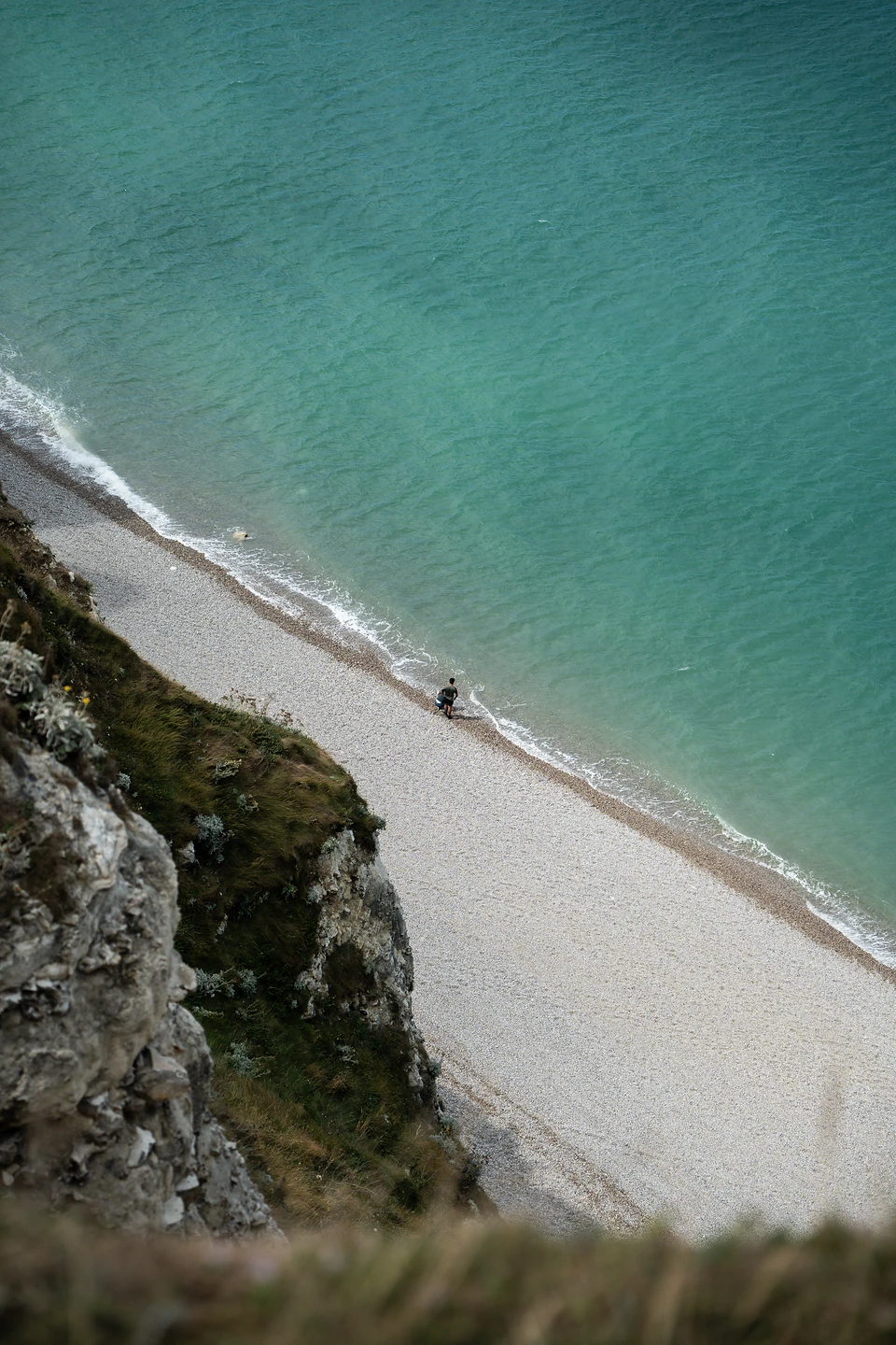 A single person at the water's edge on a pebble beach, seen from the cliff above
