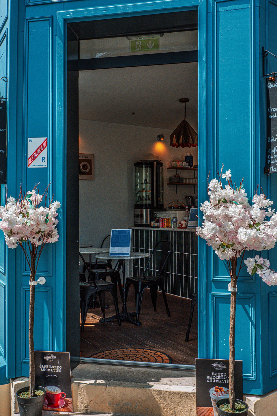 The open blue doorway of a café, flanked by two small blossom trees in pots